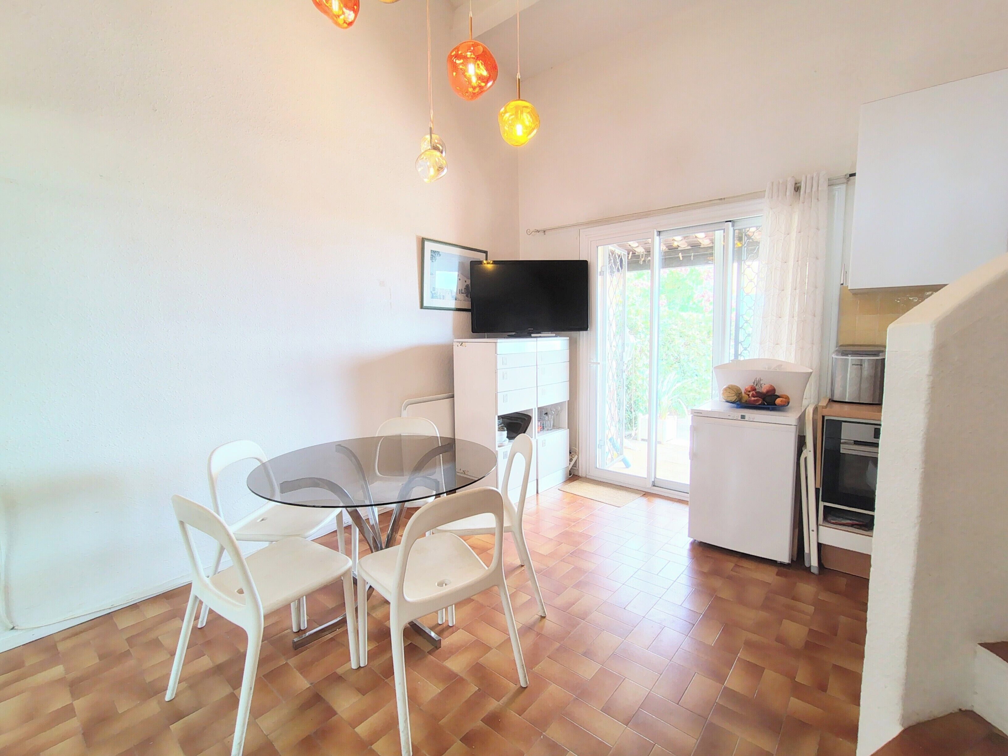 A dining area with a glass table surrounded by chairs, a television mounted on the wall, and a kitchen area with a refrigerator and a toaster.