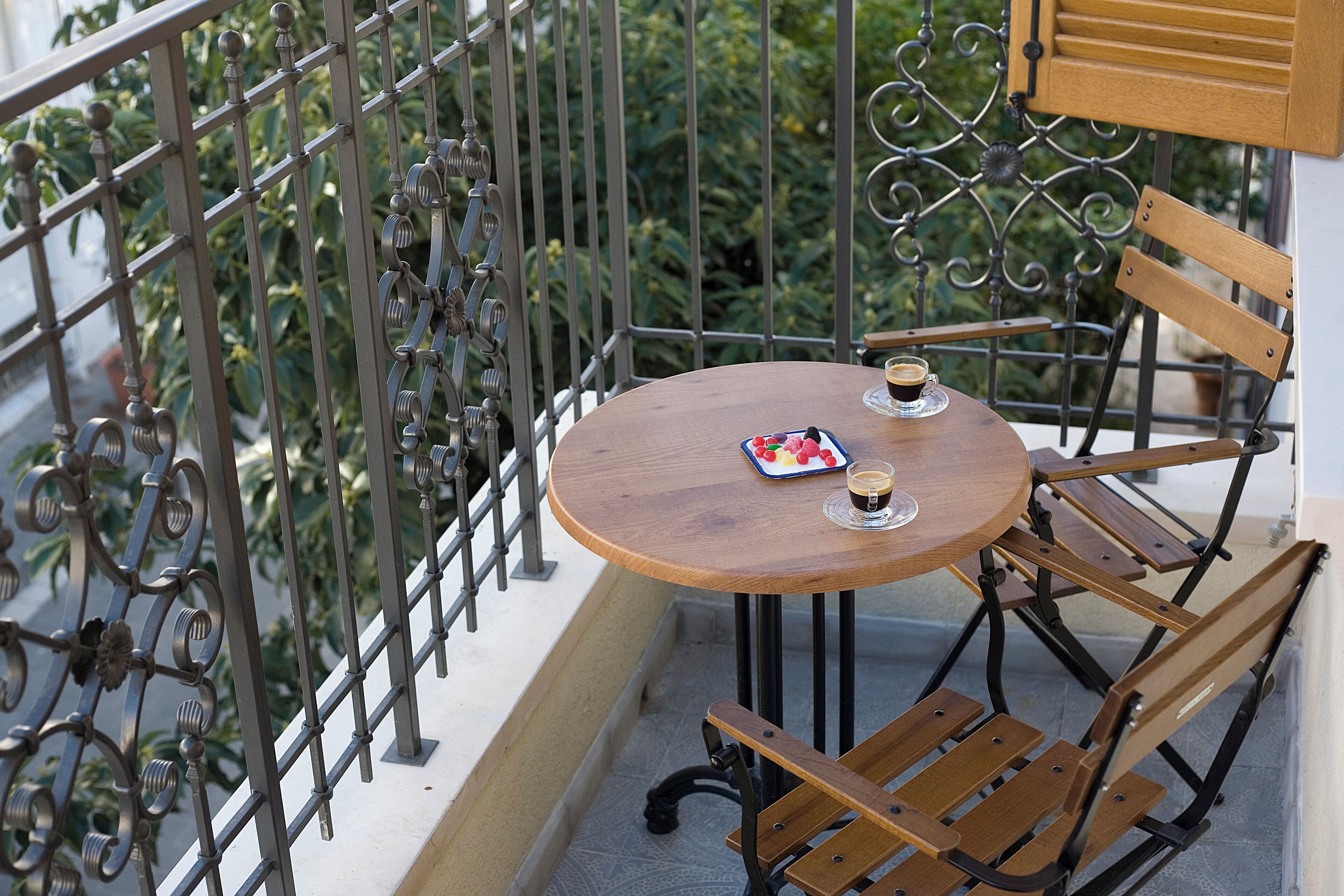 A round wooden table with two chairs, a plate of strawberries, and a cup of coffee on a balcony with metal railings and greenery in the background.