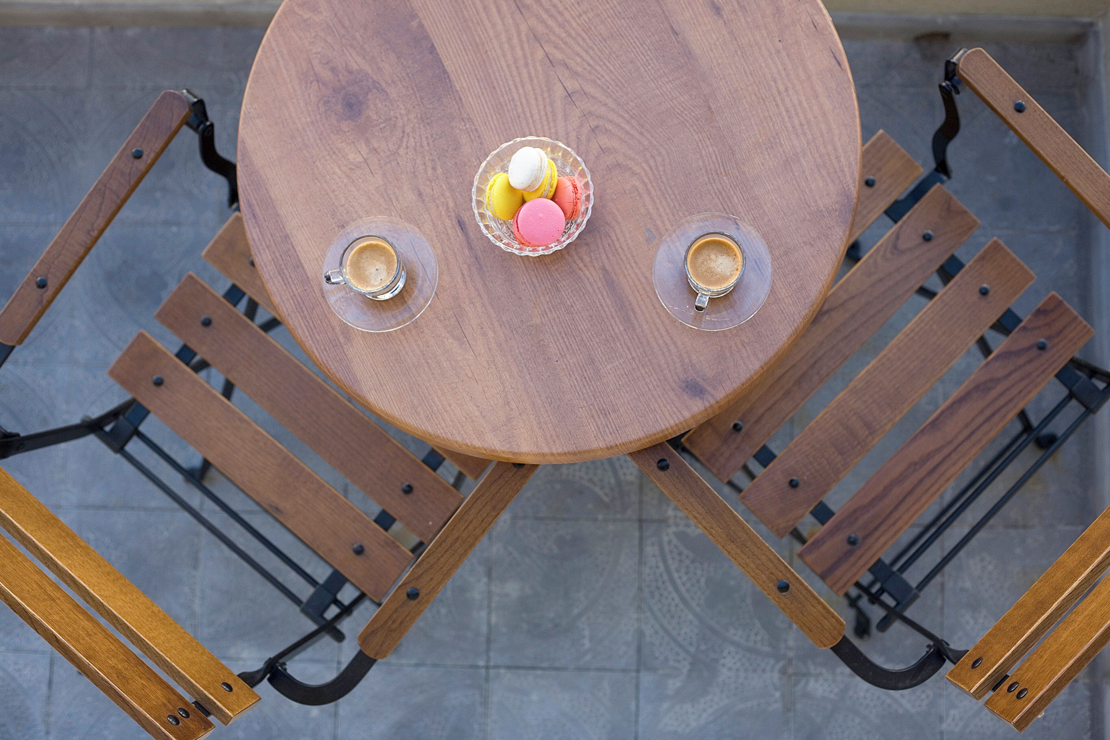 Uma mesa de madeira redonda com duas cadeiras, cada uma com uma chávena de café e uma taça de macarons coloridos.