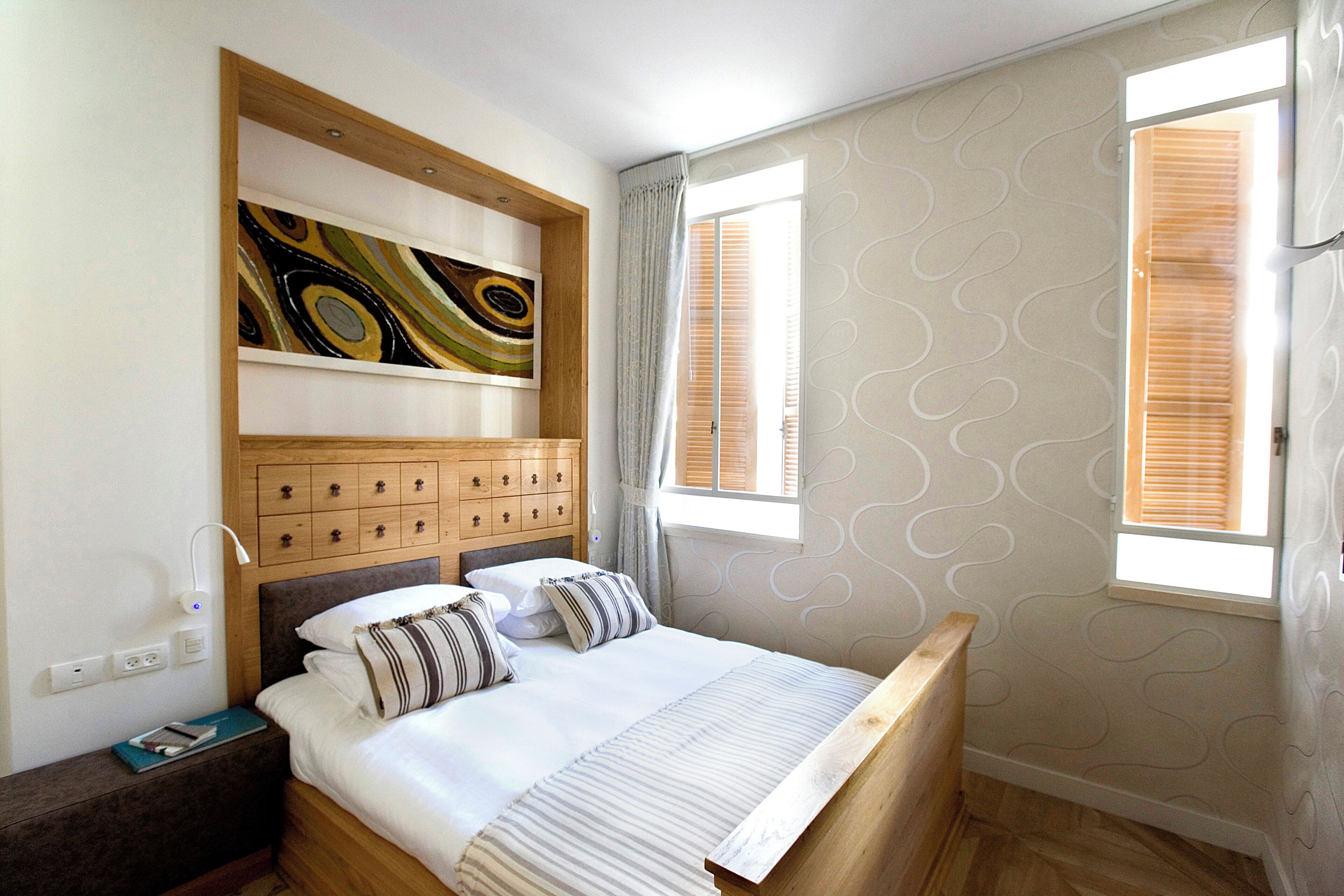 A bedroom with a wooden headboard, a bed with white bedding, a nightstand with a lamp, a wall-mounted artwork, and a window with blinds.
