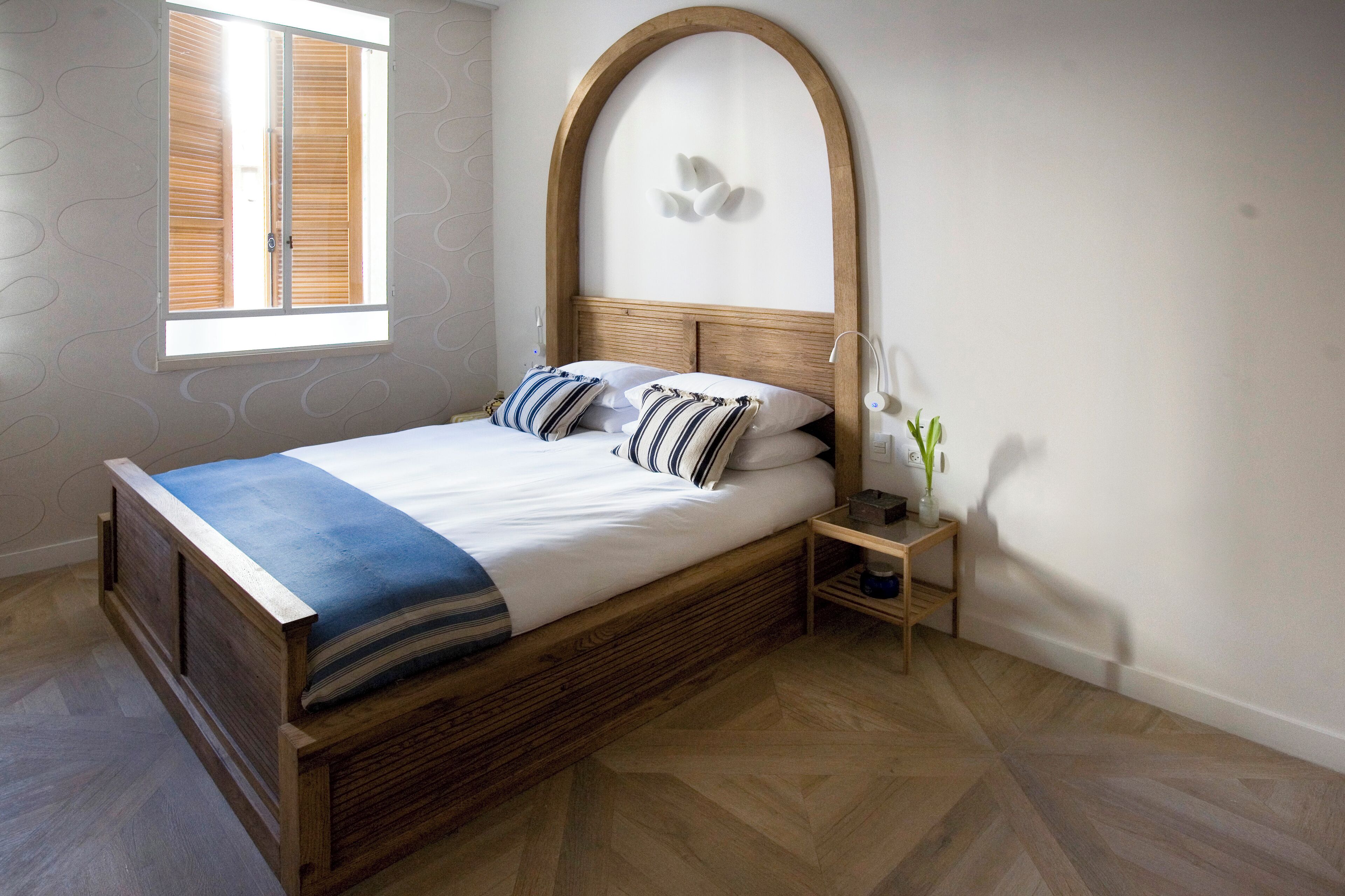 A neatly made bed with a wooden headboard, a bedside table with a lamp, and a window with blinds.