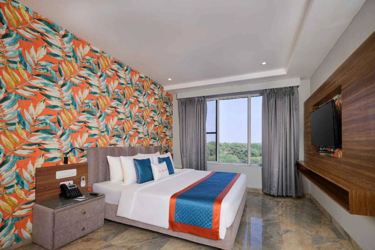 Habitación de hotel con una cama grande, una mesita de noche, un televisor montado en la pared y una ventana con cortinas.