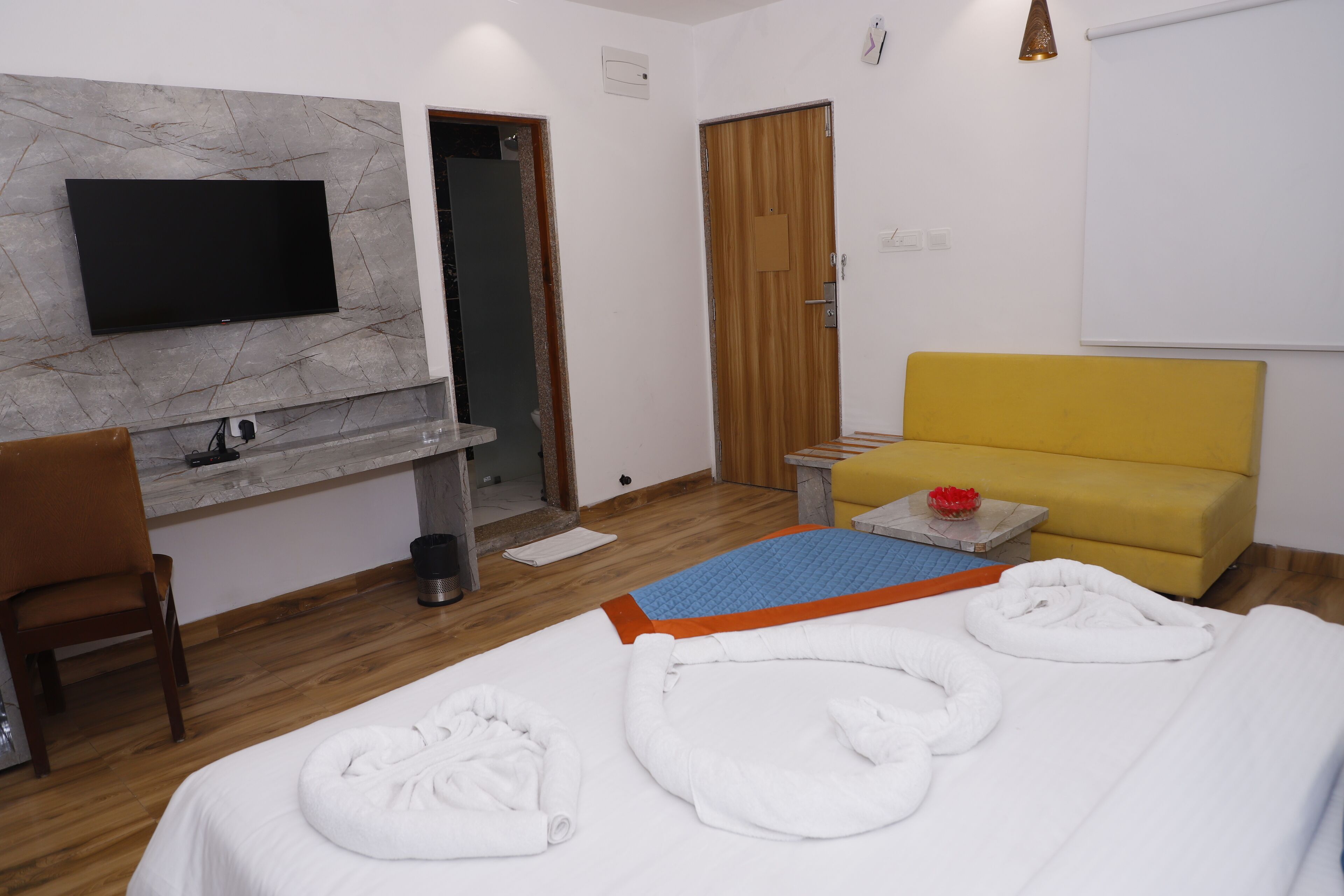 A hotel room with a bed, a yellow sofa, a TV mounted on a marble wall, a wooden door, and a small table with a red vase.