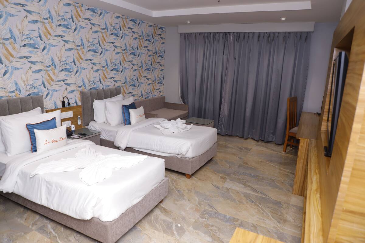 Habitación de hotel con dos camas, un ventanal grande con cortinas grises, un cabecero de madera y piso de piedra.