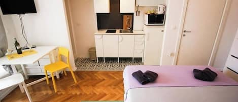 A compact hotel room with a bed, a desk with a chair, a kitchenette, and a TV mounted on the wall.