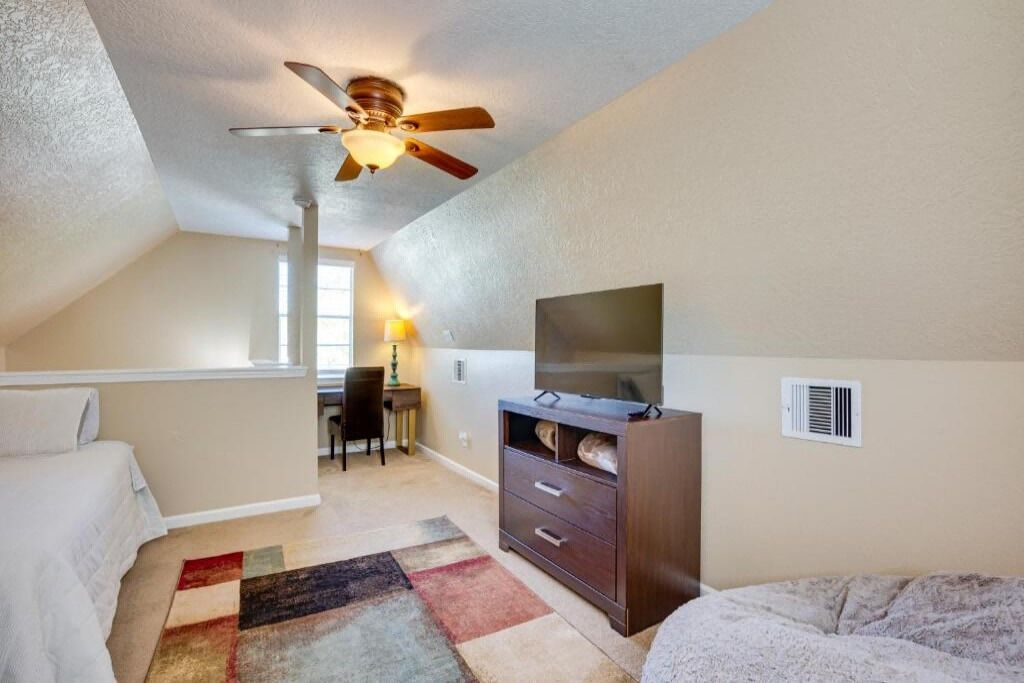 Ein Schlafzimmer mit schrägem Dach, Deckenventilator, Fernseher auf einer Holzkommode, Schreibtisch mit Lampe und einem farbenfrohen Teppich.