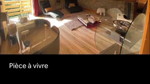 Ein gemütliches Dachzimmer mit Holzbalkendecke, Steinwand, Kamin, einer Couch, einem Esstisch und einer kleinen Küchenzeile.