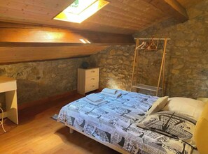 A bedroom with a stone wall, a wooden ceiling, a bed with patterned bedding, a desk, and a hanging chair.