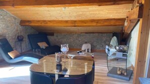 Ein gemütlicher Raum mit Holzbalkendecke, Steinwand, rundem Tisch mit Stühlen und einem Steinofen.