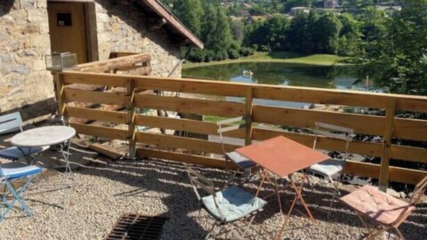 Ein rustikales Steinhaus mit einer Holzdiele, Sitzgelegenheiten im Freien und Blick auf einen See und Berge.