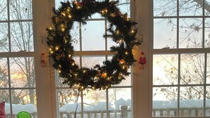 A cozy seating area with a Christmas wreath and lights, a window with a view of snow-covered trees, and two candles on a wooden table.