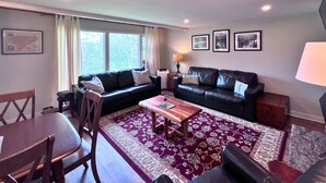 A living room with a dark leather sofa, a wooden coffee table, and a patterned rug. There are two windows with curtains, a lamp, and framed pictures on the wall. - Cozy Ski-Mountain Getaway (Sugar Mountain)