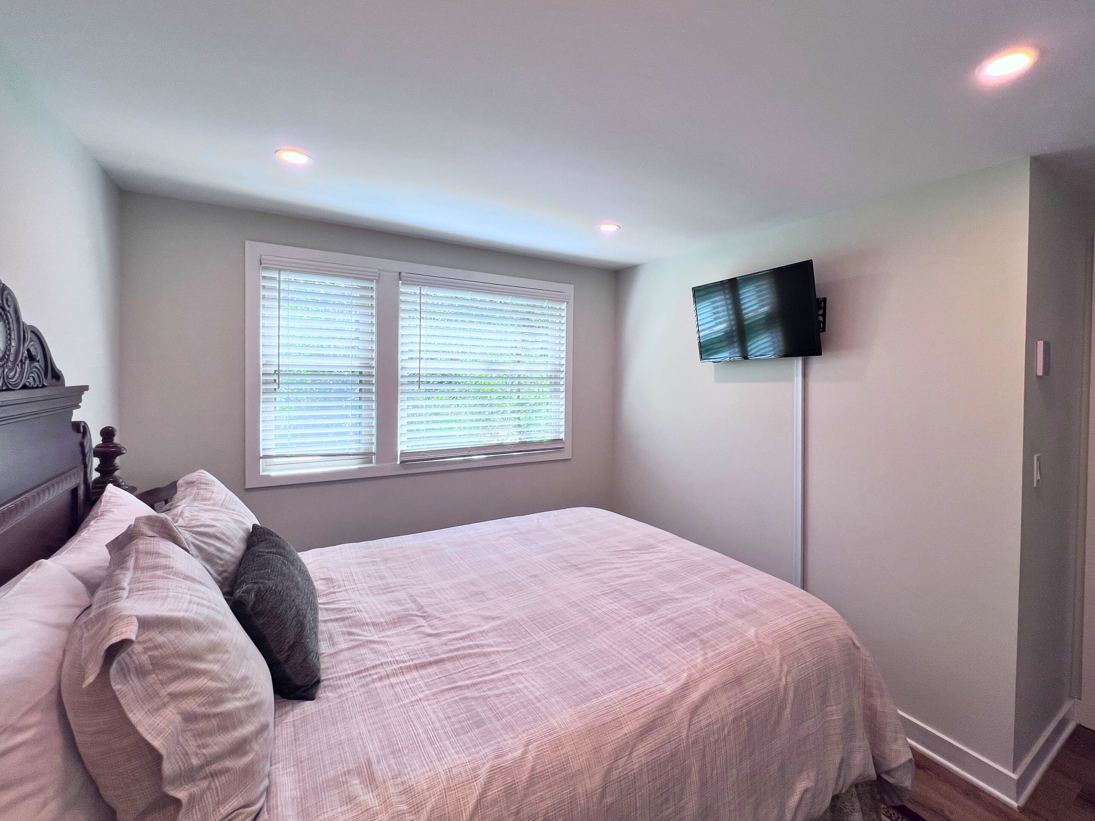 A bedroom with a bed, a television mounted on the wall, and a window with blinds.