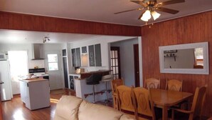 A living room with a beige sofa, wooden dining table, and chairs. There is a kitchen area with a refrigerator, stove, and bar stools. A ceiling fan is visible, and the walls are paneled.