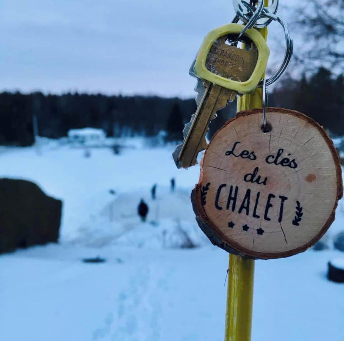 Un porte-clés en bois avec l’inscription « Les clés du CHALET » fixé à un bâtonnet jaune.