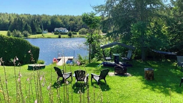 A lakeside outdoor area with chairs and a table, surrounded by greenery and a view of a house across the water.