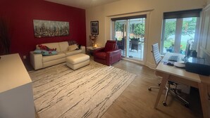 Una sala de estar con pared roja, sofá blanco, sillón rojo, puerta de vidrio a un patio y un escritorio con una computadora portátil.