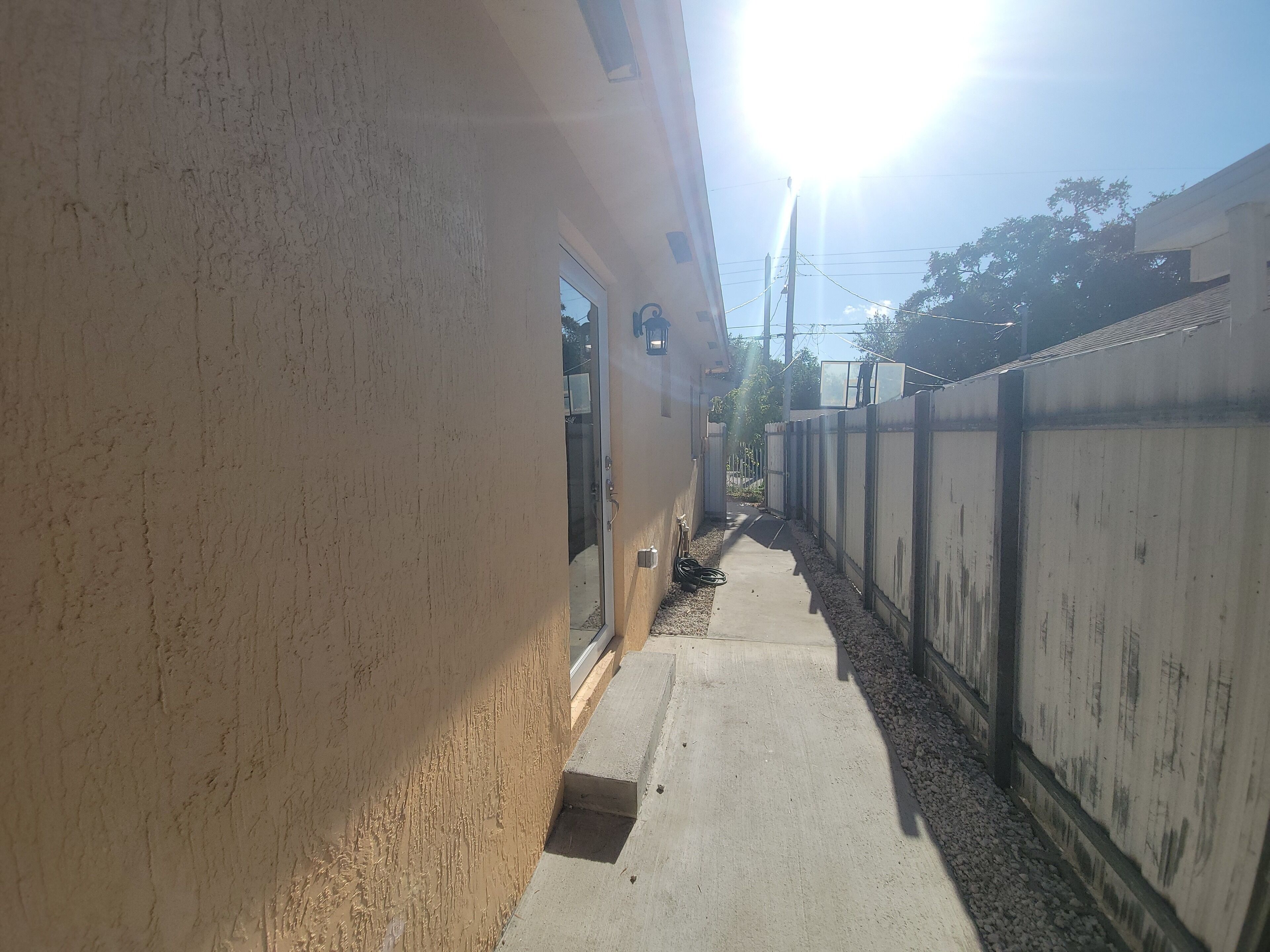 A narrow concrete pathway flanked by a beige wall and a metal fence, leading to a building entrance.