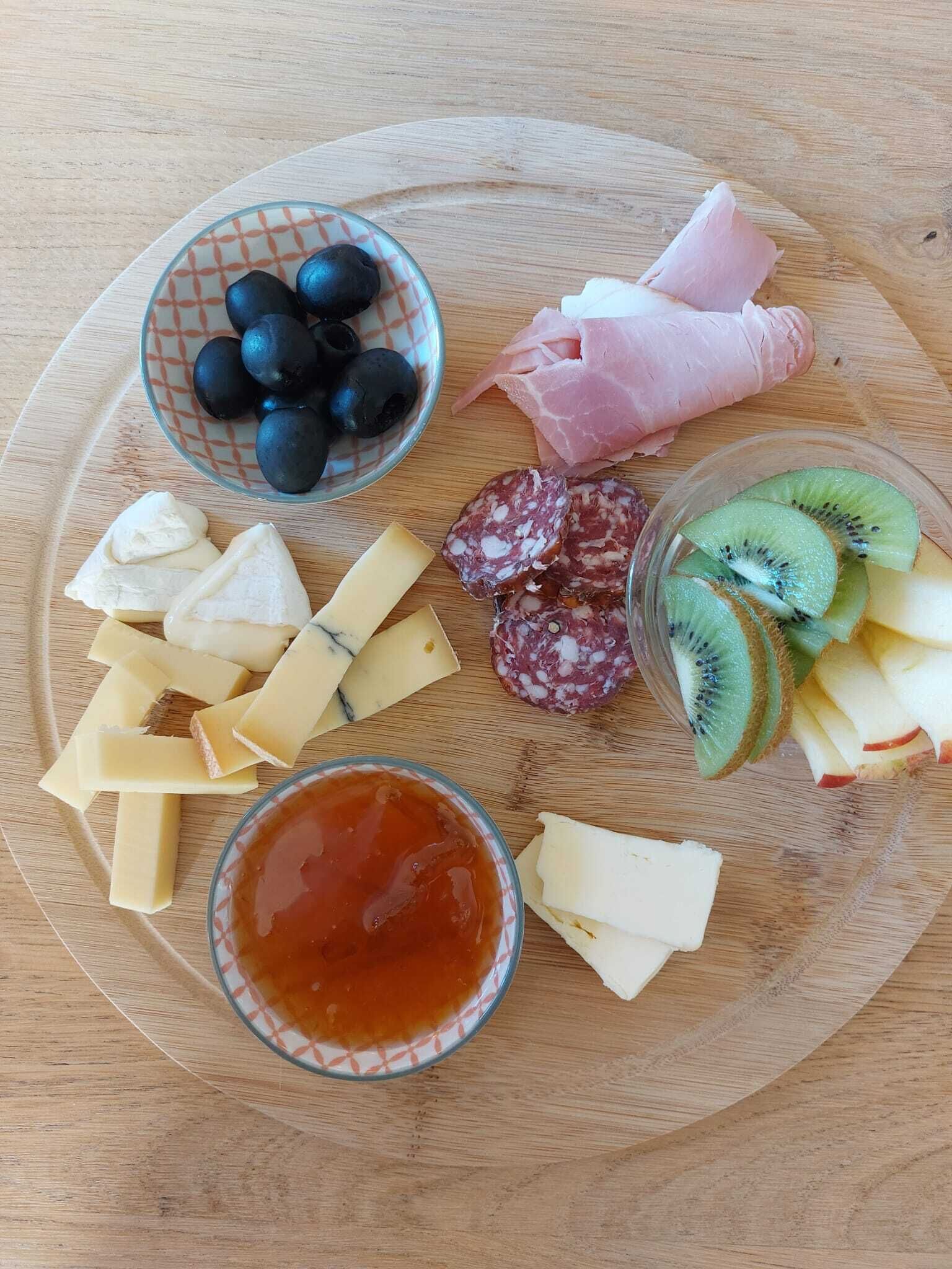 Una tabla de madera con varios quesos, embutidos, aceitunas, rodajas de kiwi y un bol de mermelada.