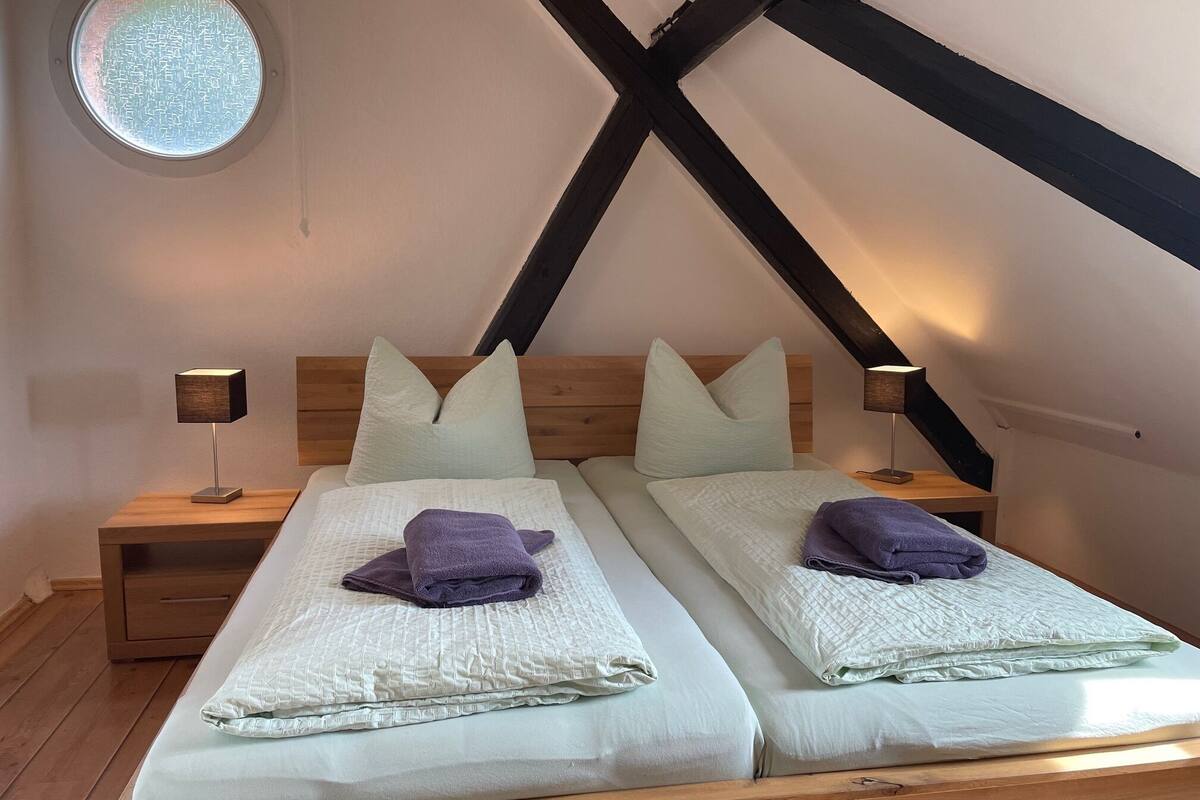 A bedroom with two beds, wooden headboards, a small bedside table with a lamp, and a circular window.