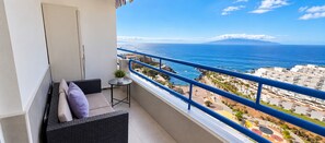 A balcony with a view of the sea and a mountain, a small table with a plant, and a sofa with cushions.
