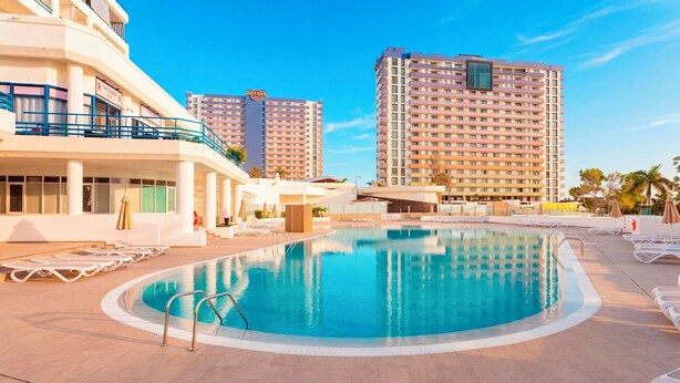 A hotel pool area with lounge chairs, a swimming pool, and high-rise buildings in the background.
