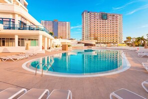 A hotel pool area with lounge chairs, a swimming pool, and high-rise buildings in the background.