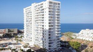 A tall, white multi-story hotel with balconies, located near a coastal area with clear skies.