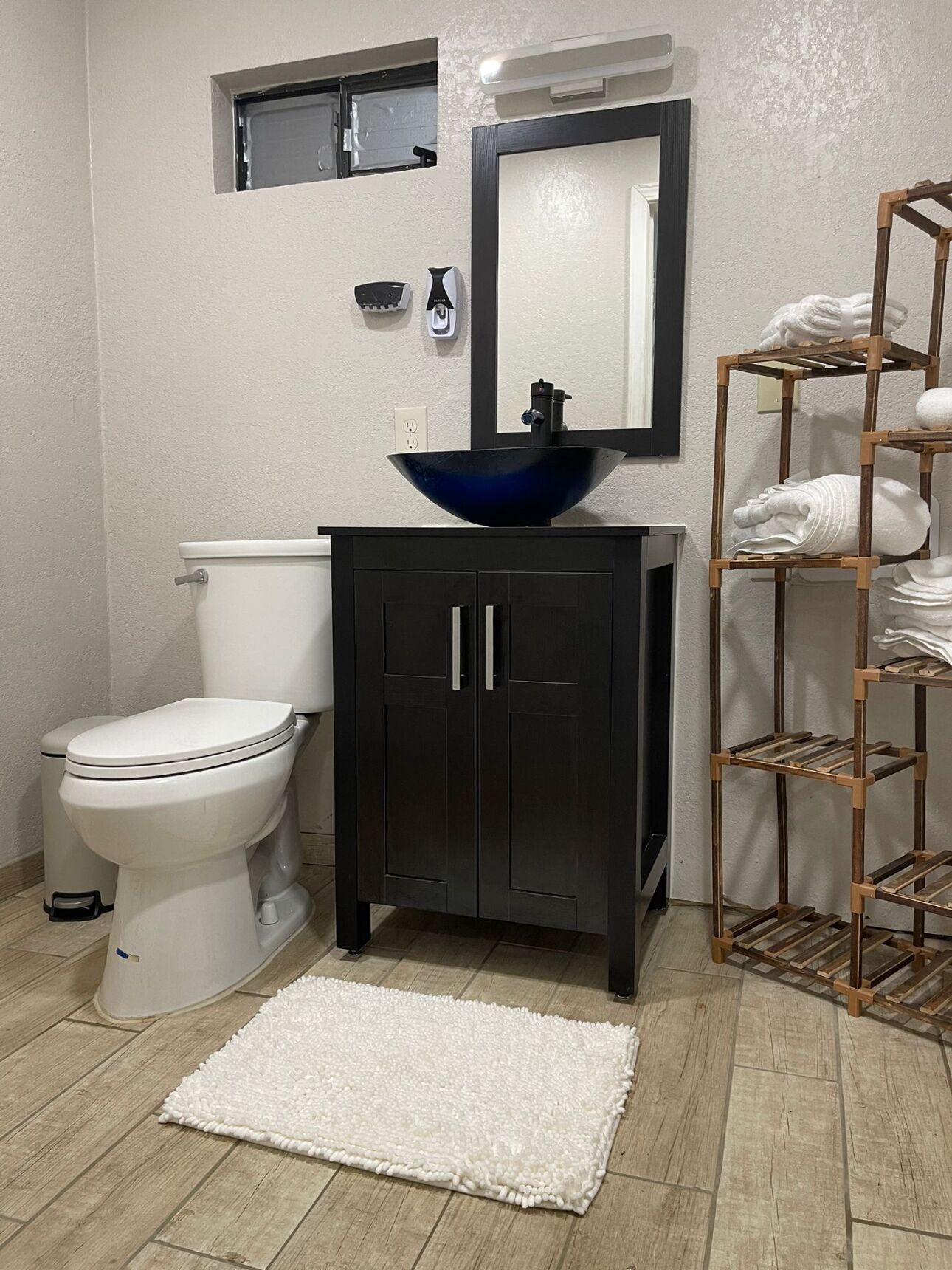 Une salle de bain avec des toilettes blanches, un meuble noir avec un lavabo bleu, un porte-serviettes en bois et un tapis blanc sur le sol.