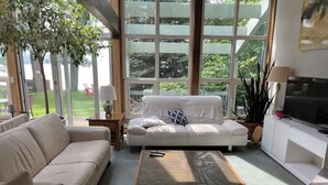 A modern living room with a white sofa, a coffee table, and a TV on a white cabinet. Large windows provide a view of the outdoors. - Magnificient view on the St-Lawrence river with private beach (Lévis)