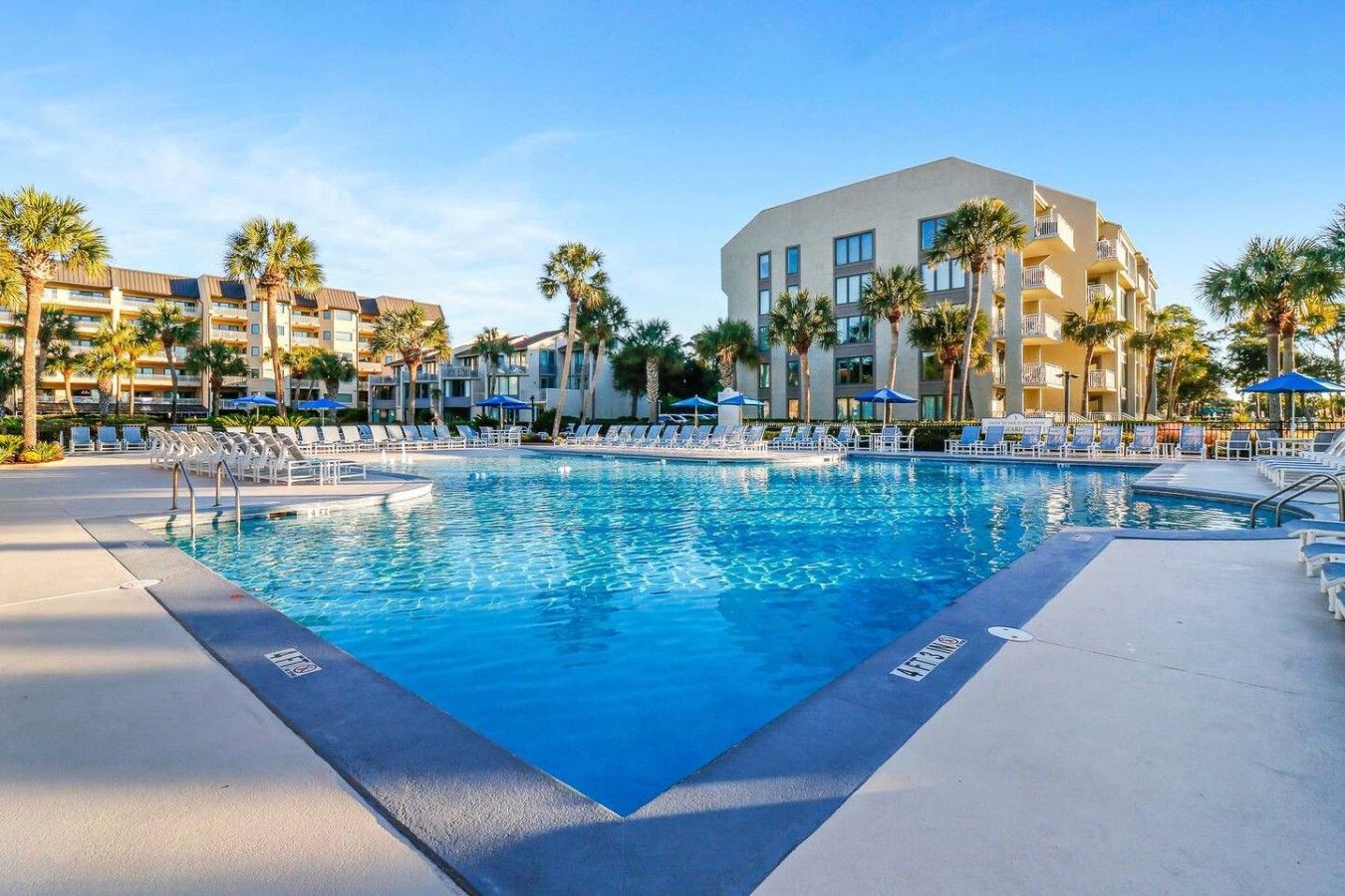 A swimming pool with lounge chairs and umbrellas, surrounded by palm trees and a hotel building.