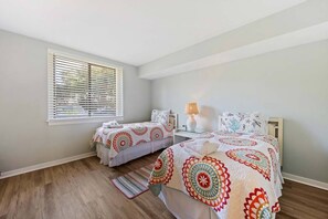 Une chambre avec deux lits, une fenêtre avec des stores, une lampe de chevet et un tapis à motifs.