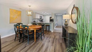 Un coin repas avec une table et des chaises en bois, une cuisine à l’arrière-plan et une horloge au mur.