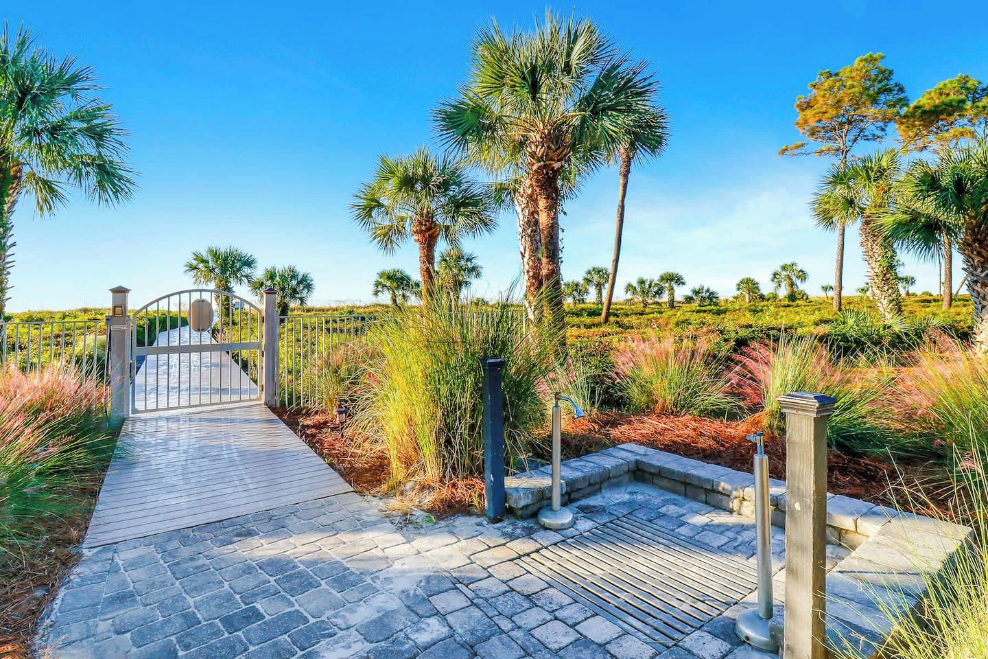 A paved pathway leading to a gated entrance, surrounded by palm trees and tropical vegetation.