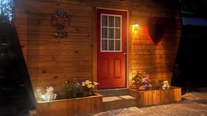 Una cabaña de madera con forma de corazón, con una puerta roja, un pequeño porche con macetas de flores y un accesorio de luz con forma de corazón sobre la puerta.