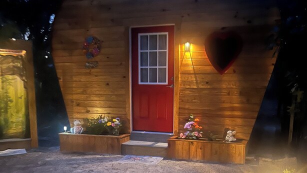 Una cabaña de madera con forma de corazón, con una puerta roja, un pequeño porche y una ventana con forma de corazón.