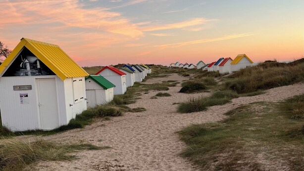 Een rij strandhutten met een zandpad er naartoe, onder een zonsondergang.