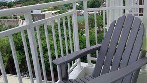 Une chaise Adirondack grise sur un balcon doté de balustrades blanches, donnant sur un quartier résidentiel verdoyant avec des bâtiments.