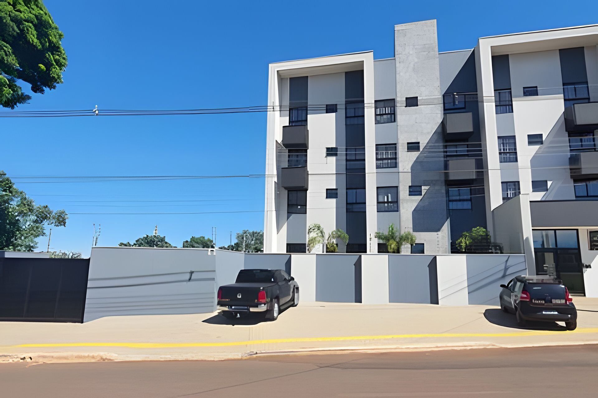 Um edifício moderno de vários andares com telhado plano, janelas amplas e um estacionamento com dois carros.