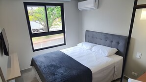 Un letto singolo con una testata grigia, una finestra vista alberi e strada, e un condizionatore d'aria montato a parete.