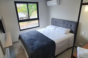 A single bed with a grey headboard, a window with a view of trees and a road, and an air conditioning unit mounted on the wall. - Residencial HS (Foz do Iguacu)