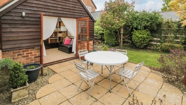 A cozy wooden cottage with a patio table and chairs, a small seating area with cushions, and a garden with various plants and a wooden fence. - Charming 3 Bed Home with Garden & Parking near Worcester (Lower Broadheath)
