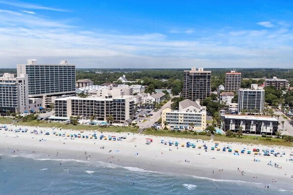 A coastal cityscape with a beachfront hotel featuring multiple floors, a beach with umbrellas, and a clear sky.