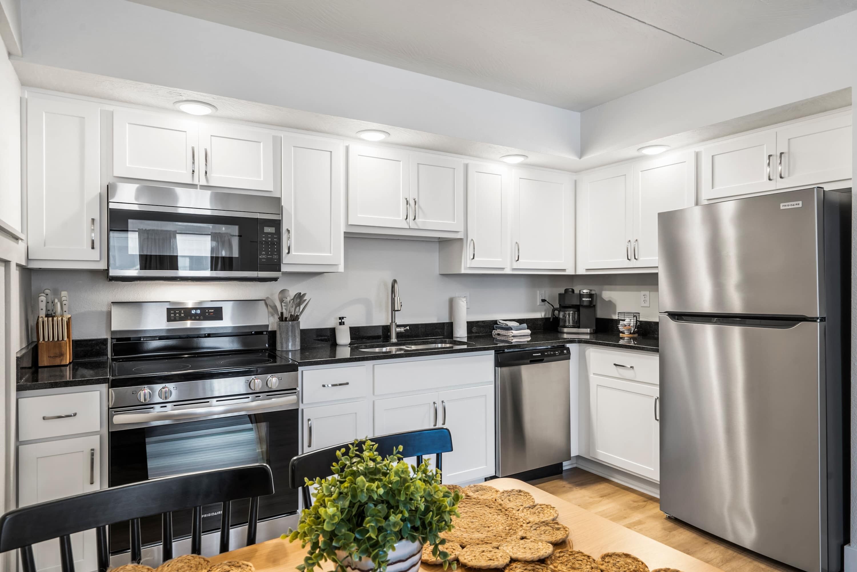 A modern kitchen with white cabinets, stainless steel appliances, and a wooden dining table with a plant and cookies.