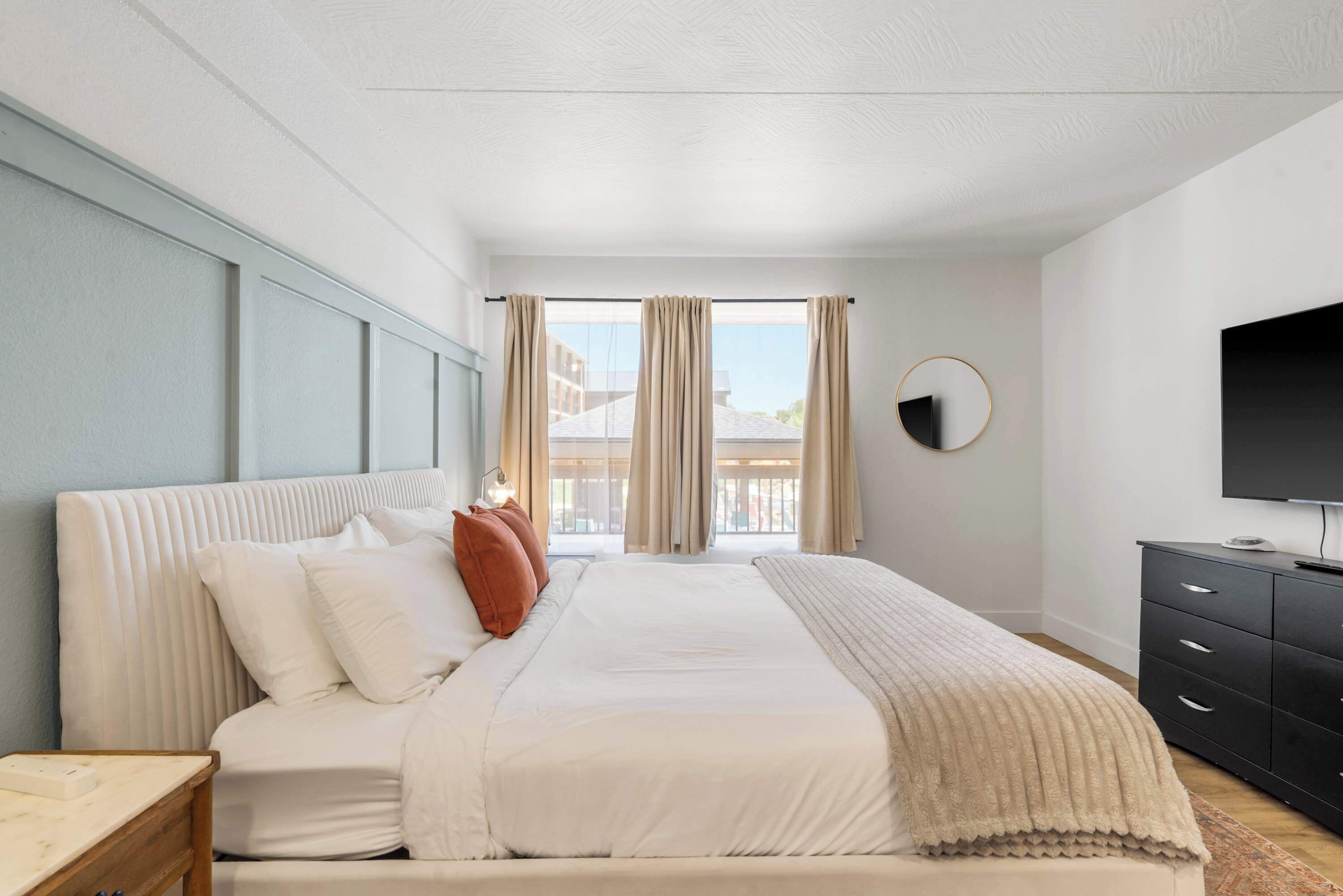 A modern bedroom with a large bed, a bedside table, a dresser, and a TV on the wall.