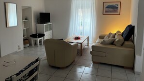En stue med sofa, sofabord, fjernsyn på et skab og et vindue med let gardin.