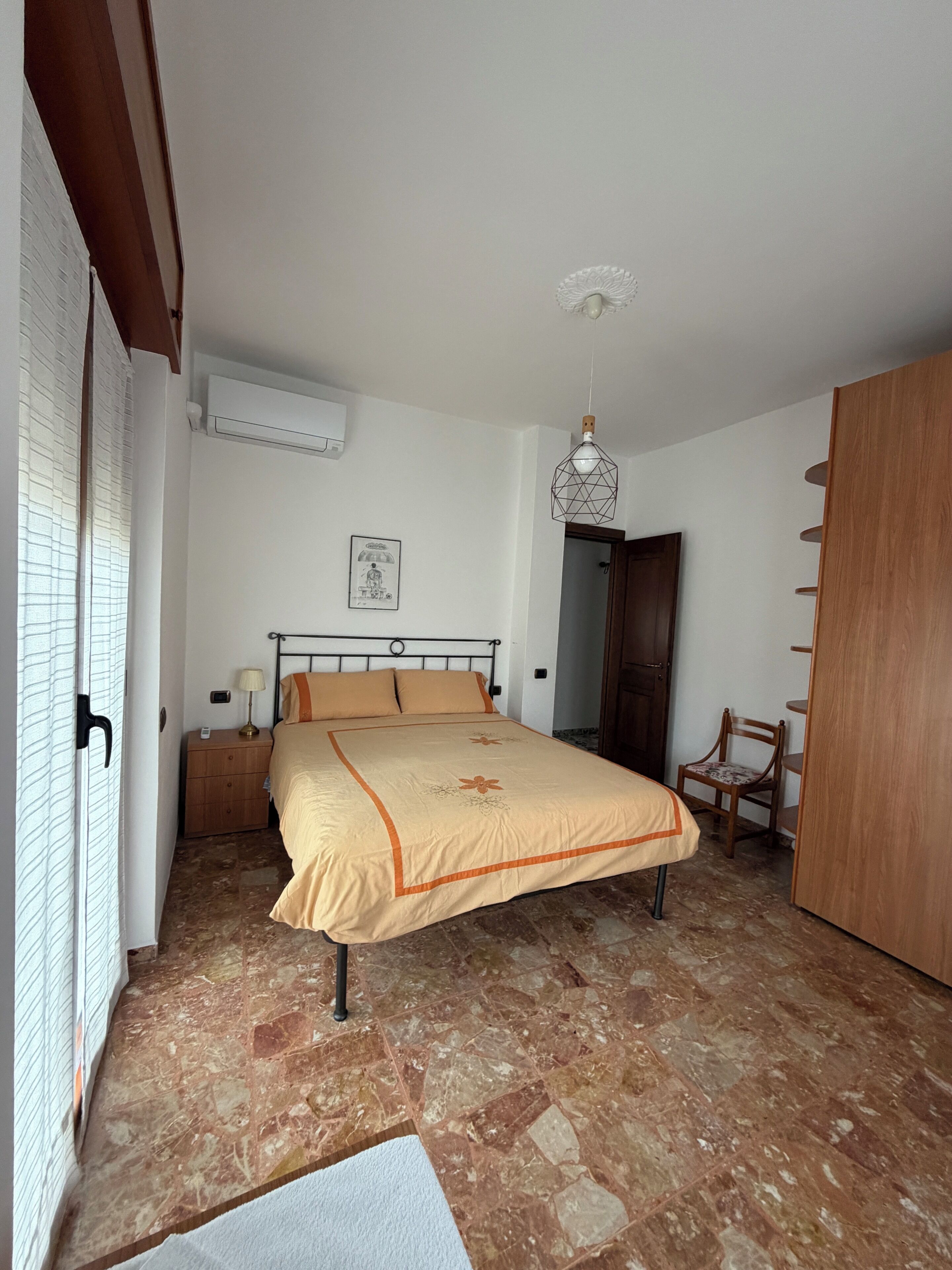 Une chambre à coucher comprenant un lit, une table de chevet, une chaise, une armoire, une porte et un climatiseur fixé au mur.