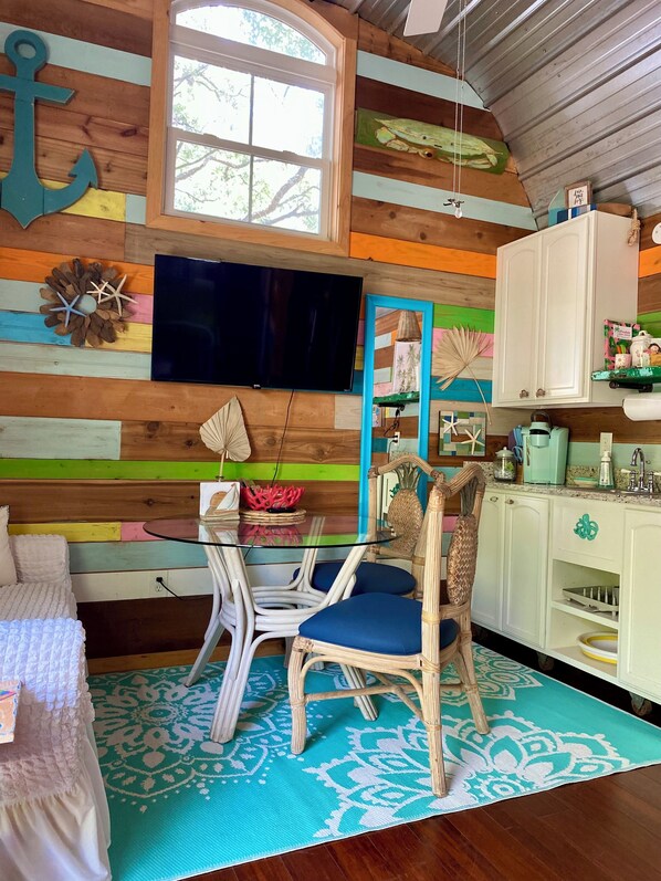A cozy cabin interior with a wooden wall, a television, a dining table with chairs, a bed, and a kitchen area.