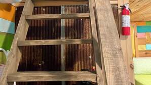 A wooden ladder leading to a lofted sleeping area with a corrugated metal ceiling and a fire extinguisher mounted on the wall. - Perdido Surf Shack-tiny home on a hobby farm close to Perdido Key Beach and NAS (Pensacola)