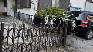 Un immeuble résidentiel doté d’une entrée sécurisée par un portail, une voiture noire garée devant, et un petit jardin agrémenté de fleurs blanches.